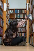 étudiant fille, séance sur le sol dans le bibliothèque et choisir livre pour connaissance, apprentissage ou Littérature à livre magasin. femelle étudiant séance sur sol par étagère à livres dans étude, recherche ou éducation photo