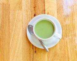 Haut vue de chaud condensé Lait matcha vert thé servi dans blanc tasse et petit assiette avec petit thé cuillère sur marron réveillé Contexte photo