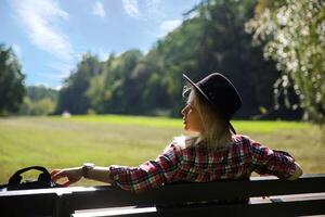 Jeune femme dans une chapeau et une plaid chemise est assis sur une banc près le prairie. flou Contexte. sélectif se concentrer. haute qualité photo