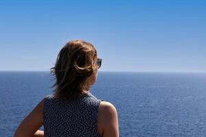 une Jeune femme dans des lunettes de soleil regards dans le distance contre le Contexte de le mer et montagnes. haute qualité photo