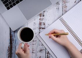 Jeune femme travail avec une portable et en buvant café pendant isolement ou quarantaine. proche est un vide carnet et stylo. vue de au-dessus de. travail espace. haute qualité photo