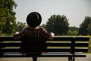 Jeune femme dans une chapeau et une plaid chemise est assis sur une banc près une Prairie dans le parc.fond flou. sélectif se concentrer. haute qualité photo