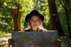 Jeune caucasien femme dans une chapeau à la recherche à une géographique carte dans le ombre de des arbres dans une parc ou forêt. haute qualité photo