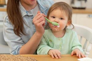 content famille à maison. mère alimentation sa bébé fille de cuillère dans cuisine. peu bambin enfant avec désordonné marrant visage mange en bonne santé nourriture à maison. Jeune femme maman donnant nourriture à enfant fille photo