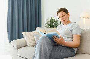 portrait de une content mature femme relaxant sur canapé et en train de lire une livre dans une moderne maison photo