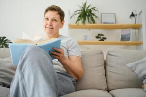 portrait de une content mature femme relaxant sur canapé et en train de lire une livre dans une moderne maison photo