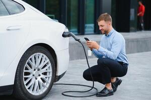 homme mise en charge le sien électrique voiture à charge station et en utilisant téléphone intelligent photo