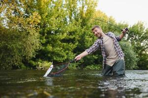 homme avec pêche canne à pêche, pêcheur Hommes dans rivière l'eau Extérieur. contagieux truite poisson dans filet. été pêche loisir photo