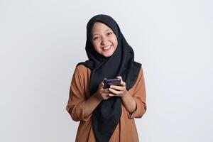 excité magnifique asiatique femme dans marron chemise et hijab en utilisant mobile téléphone, célébrer succès, avoir bien nouvelles isolé sur blanc Contexte photo