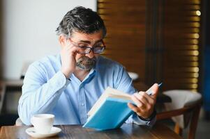 Sénior vieux homme en train de lire une livre dans une café boutique, profiter le sien littéraire loisir photo