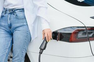 content Jeune adulte femme souriant large, à la recherche loin, mise en charge voiture batterie de petit Publique gare, permanent près électrique auto. photo