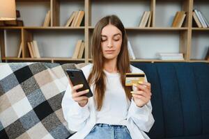 content adolescent Faire achats en ligne séance sur canapé à maison. Jeune femme insérer crédit carte nombre sur site Internet avec sa intelligent téléphone. photo