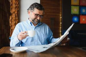 fermer portrait de sérieux Sénior Beau homme en train de lire journal, ayant café Pause et séance à tableau. photo