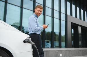 homme mise en charge le sien électrique voiture à charge station et en utilisant téléphone intelligent photo