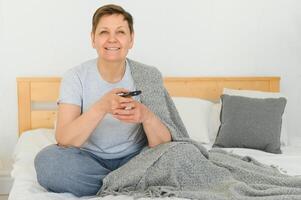 âge moyen femme séance sur le canapé et tournant sur le la télé avec une éloigné contrôle. photo