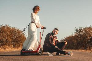 couple de mariage souriant chevauchant un scooter le long de la route à l'extérieur de la ville au coucher du soleil. photo