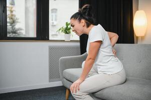 dérangé mature femme Souffrance de mal au dos, malheureux Sénior blond séance sur une canapé à vivant chambre, sentiment malaise car de douleur dans retour photo