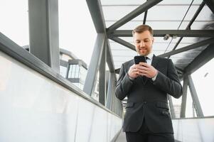 content souriant homme d'affaire portant noir costume et en utilisant moderne téléphone intelligent près Bureau à de bonne heure matin, réussi employeur à faire une traiter tandis que permanent près gratte-ciel Bureau à nuit, éclater lumière photo