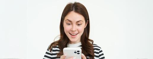 portrait de Jeune femme à la recherche à téléphone intelligent écran avec surpris, étonné visage expression, en train de lire génial nouvelles sur téléphone photo