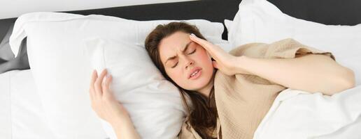 proche en haut portrait de malade femme mensonge dans sa lit et sentiment indisposé, émouvant diriger, a mal de tête, à la recherche frustré de migraine, portant pyjamas photo