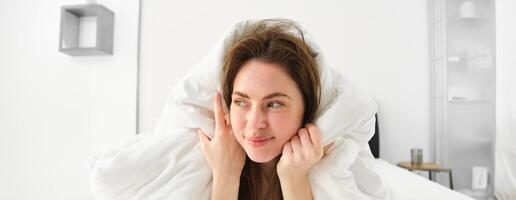 mignonne fille avec désordonné cheveux, mensonge dans lit couvert dans blanc feuilles couette, souriant et en riant coquette, dépenses temps dans sa chambre photo