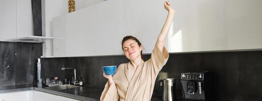 portrait de insouciant brunette femme, dansant et élongation mains dans le cuisine, en buvant café, souriant heureux dans le Matin photo