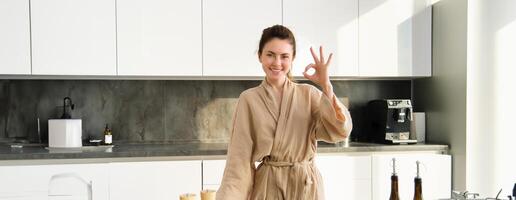 portrait de souriant brunette femme cuisine en bonne santé dîner, montrant d'accord signe, épouse permanent dans peignoir de bain dans cuisine, en train de préparer nourriture, couper des légumes sur planche photo