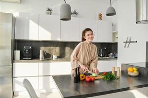 portrait de magnifique brunette femme dans le cuisine, portant peignoir de bain, couper des légumes sur conseil, cuisine en bonne santé végétarien nourriture, en train de préparer salade, fabrication une repas photo