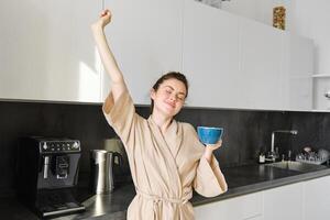 portrait de insouciant brunette femme, dansant et élongation mains dans le cuisine, en buvant café, souriant heureux dans le Matin photo