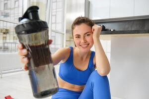 Jeune positif fille Est-ce que faire des exercices de maison, donne vous l'eau bouteille à boisson pendant formation des exercices, est assis sur yoga tapis sol dans vivant pièce photo