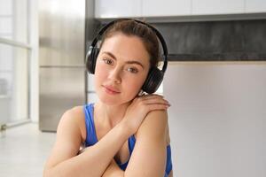 proche en haut portrait de brunette aptitude fille, écoute la musique dans sans fil écouteurs, sourit à caméra, Est-ce que faire des exercices seul à maison, est assis sur sol et des exercices photo