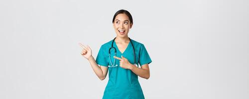 COVID-19 [feminine, soins de santé ouvriers, pandémie concept. optimiste souriant femelle asiatique infirmière, médecin dans gommages, à la recherche et montrer du doigt plus haut la gauche coin, montrant bannière, démontrer annonce photo