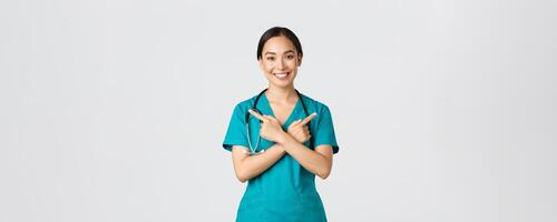 COVID-19 [feminine, soins de santé ouvriers, pandémie concept. souriant magnifique asiatique infirmière, interne ou médecin dans gommages montrer du doigt de côté et à la recherche à caméra, montrant variantes, deux les choix, blanc Contexte photo