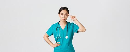 COVID-19 [feminine, soins de santé ouvriers, pandémie concept. agacé sceptique asiatique femelle infirmière ou médecin réprimande Quelqu'un agissant fou ou stupide, rouleau doigt plus de temple avec jugement visage photo