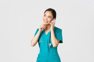 COVID-19 [feminine, soins de santé ouvriers et empêcher virus concept. portrait de souriant asiatique femelle médecin, interne dans gommages parlant sur téléphone et à la recherche réfléchi, en pensant ou fabrication choix photo