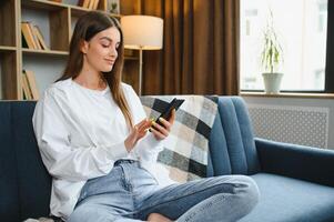 Jeune content caucasien femme elle Années 20 dans décontractée vêtements tenir dans main utilisation mobile cellule téléphone asseoir sur bleu canapé intérieur du repos à Accueil dans posséder pièce appartement. gens mode de vie concept photo