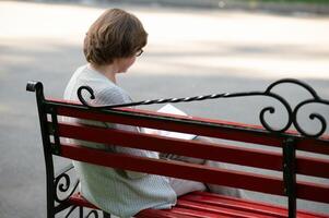 élégant personnes âgées femme dans le chemise est séance sur le banc dans une parc sur une chaud journée photo