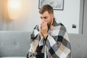 une malade homme est assis à Accueil sur une gris canapé avec une couverture. maladie, protection, corona virus, maladie, grippe photo