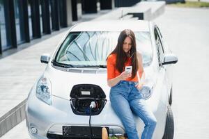 en utilisant téléphone intelligent tandis que en attendant. femme sur le électrique voitures charge station à jour. marque Nouveau véhicule. photo