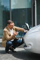 beau barbu homme séance près le sien Nouveau moderne électrique voiture et en portant prise de courant de le chargeur, tandis que voiture est mise en charge à le mise en charge station photo