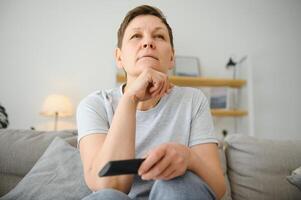 âge moyen femme séance sur le canapé et tournant sur le la télé avec une éloigné contrôle. photo