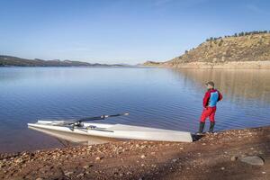 Sénior Masculin rameur et une côtier aviron coquille est atterrissage sur une rocheux rive de charretier Lac dans tomber ou hiver paysage dans nord Colorado. photo