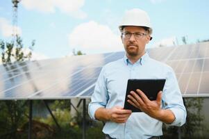 portrait de Masculin ingénieur avec tablette dans le sien mains près le solaire panneaux gare, portant casque à ensoleillé journée. vert écologique Puissance énergie génération. photo