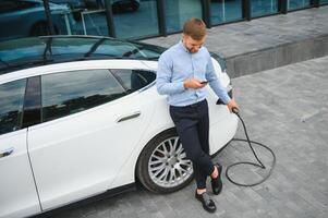 portrait de Jeune Beau barbu homme dans décontractée porter, permanent à le mise en charge station et en portant une prise de courant de le chargeur pour un électrique auto. éco électrique voiture concept. photo