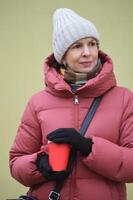 une mignonne caucasien femme dans une tricoté chapeau et hiver vers le bas veste détient une rouge papier tasse de café dans sa mains. photo