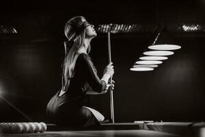 une fille avec une bandeau et une queue dans sa mains est séance sur une table dans une billard club.russe billard. noir et blanc photo