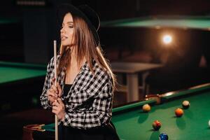 une fille dans une chapeau dans une billard club avec une queue dans sa mains.piscine Jeu photo