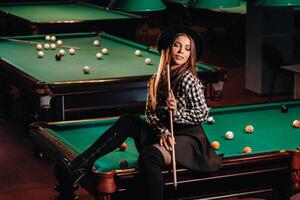 une fille dans une chapeau dans une billard club est assis sur une billard table avec une queue dans sa mains.jouer billard photo