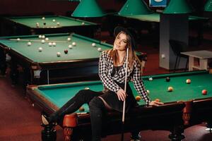 une fille dans une chapeau dans une billard club est assis sur une billard table avec une queue dans sa mains.jouer billard photo