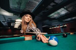 une fille dans une chapeau dans une billard club avec une queue dans sa mains les coups une jouer au ballon bassin photo